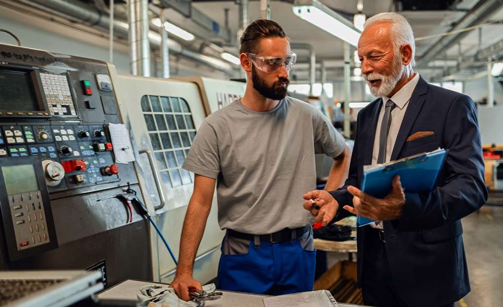 A machine operator and an older businessman have a conversation on the shop floor.