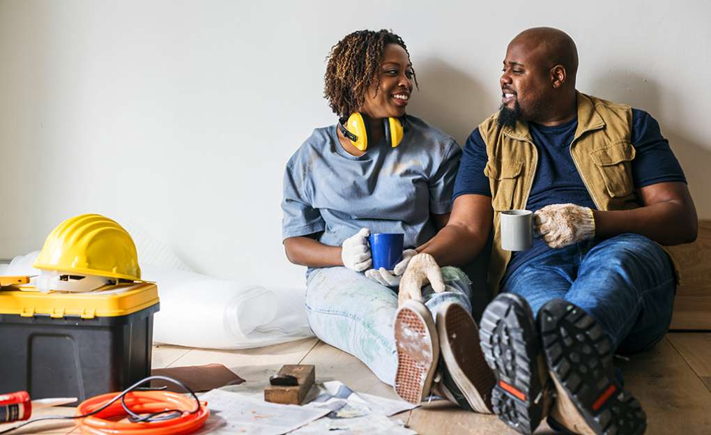 A couple sits on the floor drinking coffee in a room that is in the process of being remodeled. 