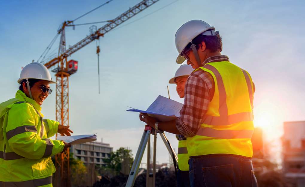 Two workers wearing hardhats review information in a notebook. A crane can be seen in the background.