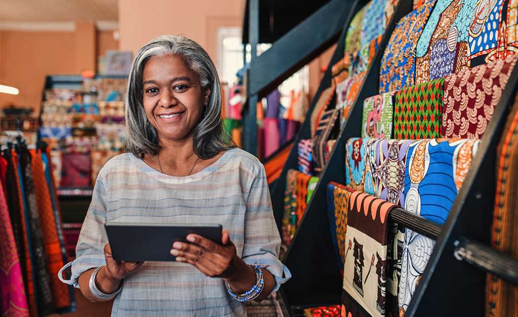 An older woman in a textile shop holds a tablet in her hands and smiles.