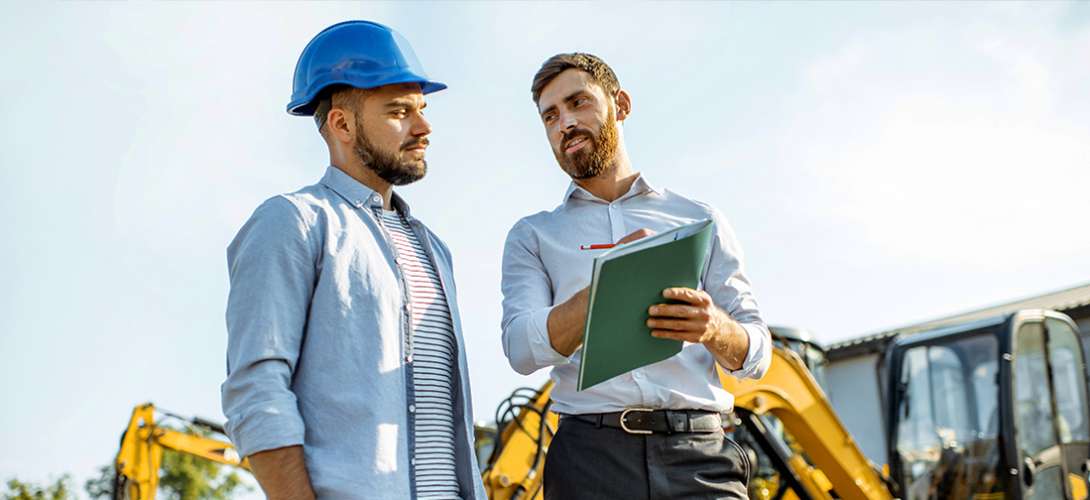 A man in a hard hat conversing with a well dressed man with a clipboard/folder. 