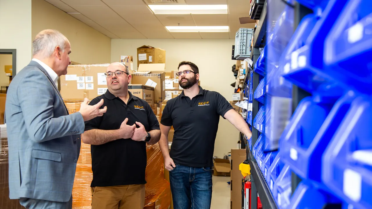 A North Easton Savings Bank lender standing and talking to two local business partners inside a storage room. To their right, shelves are filled with various blue plastic parts containers.