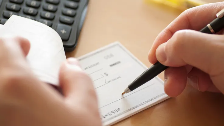 Hands signing a check with a pen at a desk with a calculator in the background