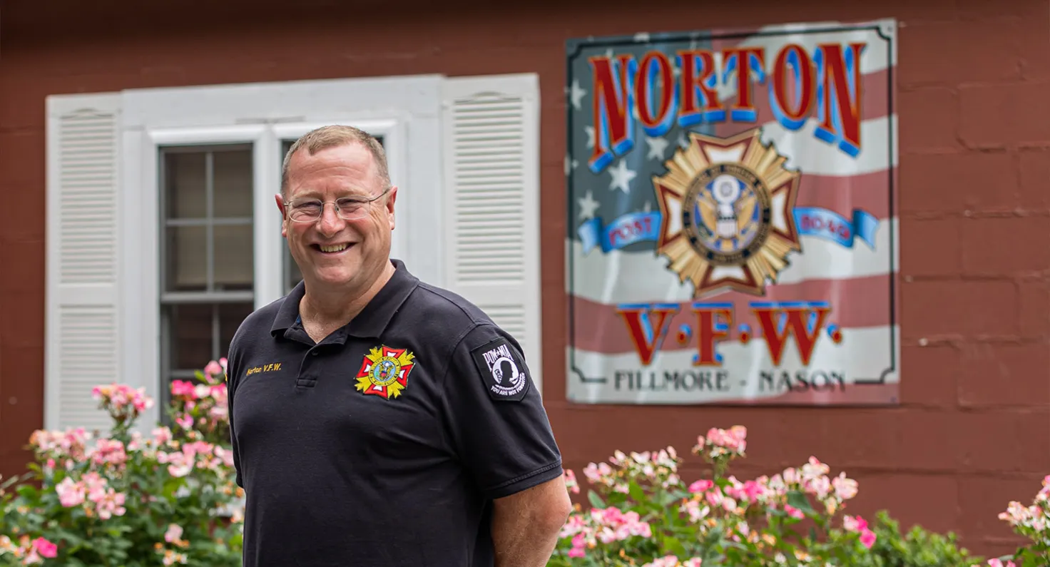 A veteran smiling in front of a red building with a sign that reads Norton VFW.